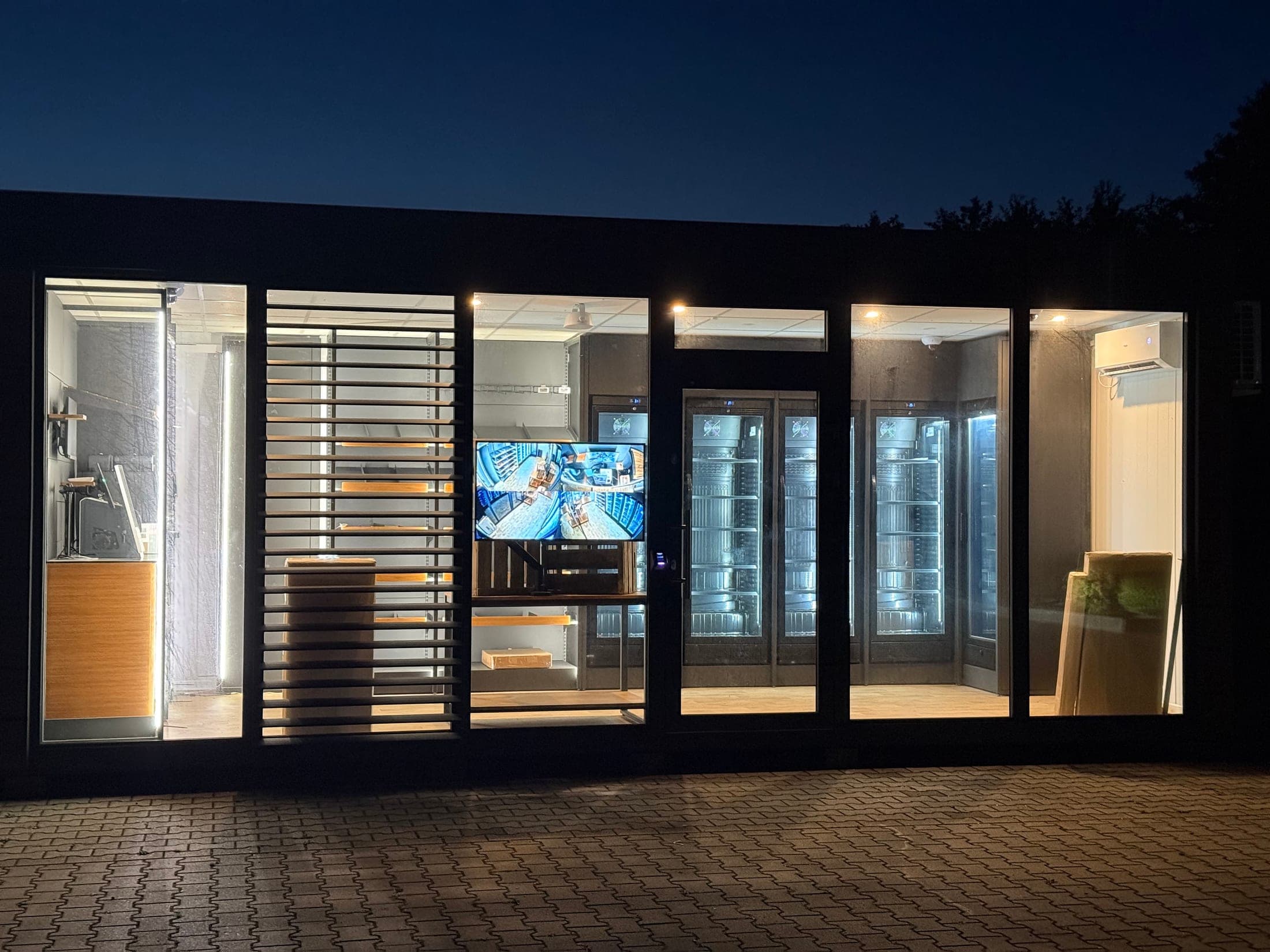 Die fertige Glasfront des Shop-Containers bei Nacht — Kühlgeräte, Regale und der Kamera-Monitor sind beleuchtet