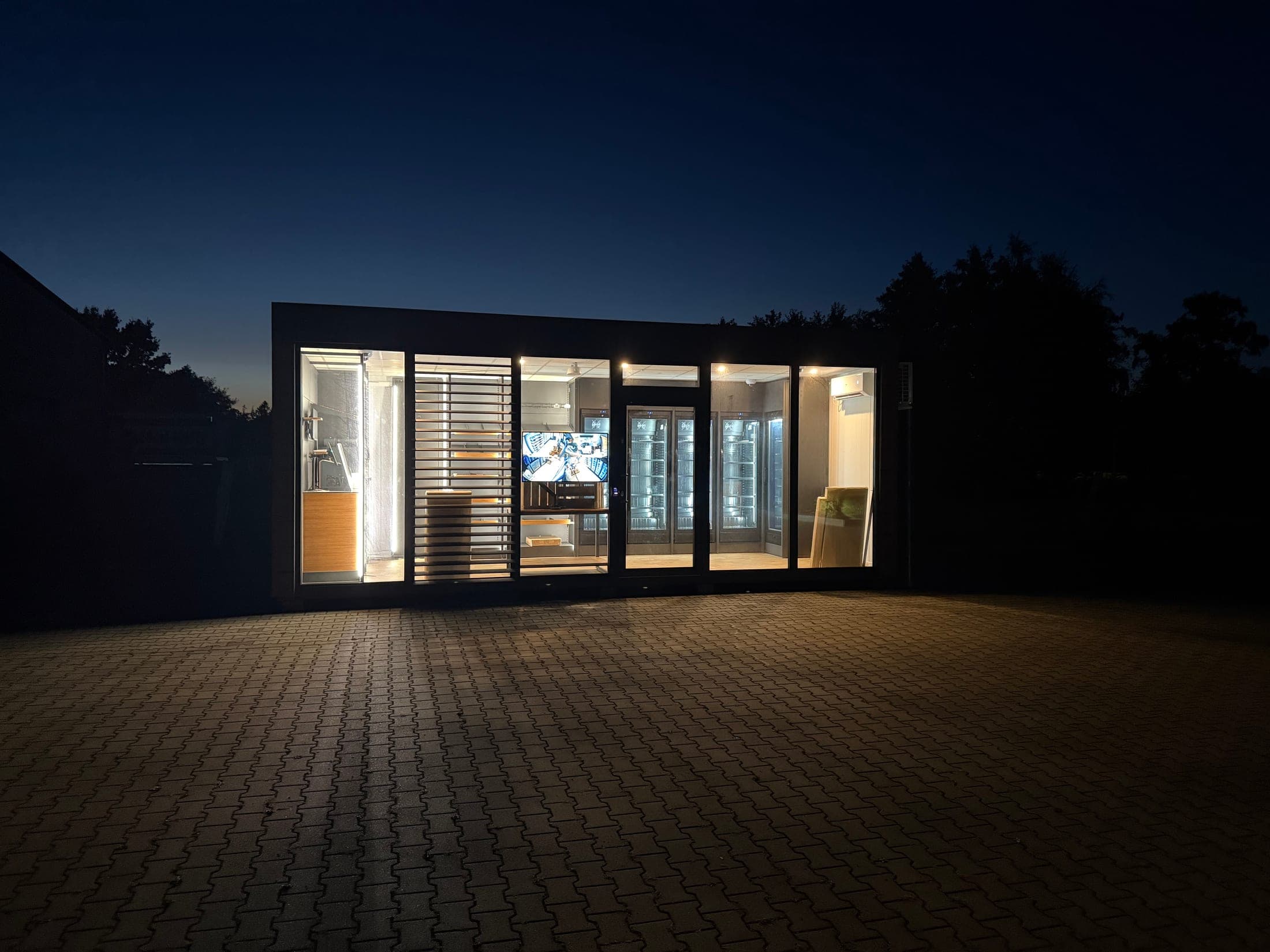Der neue 24/7-Shop-Container von Albert Wilts in Upgant-Schott bei Nacht — beleuchtete Glasfront mit Blick auf Kühlgeräte und den Live-Monitor des Kamerasystems