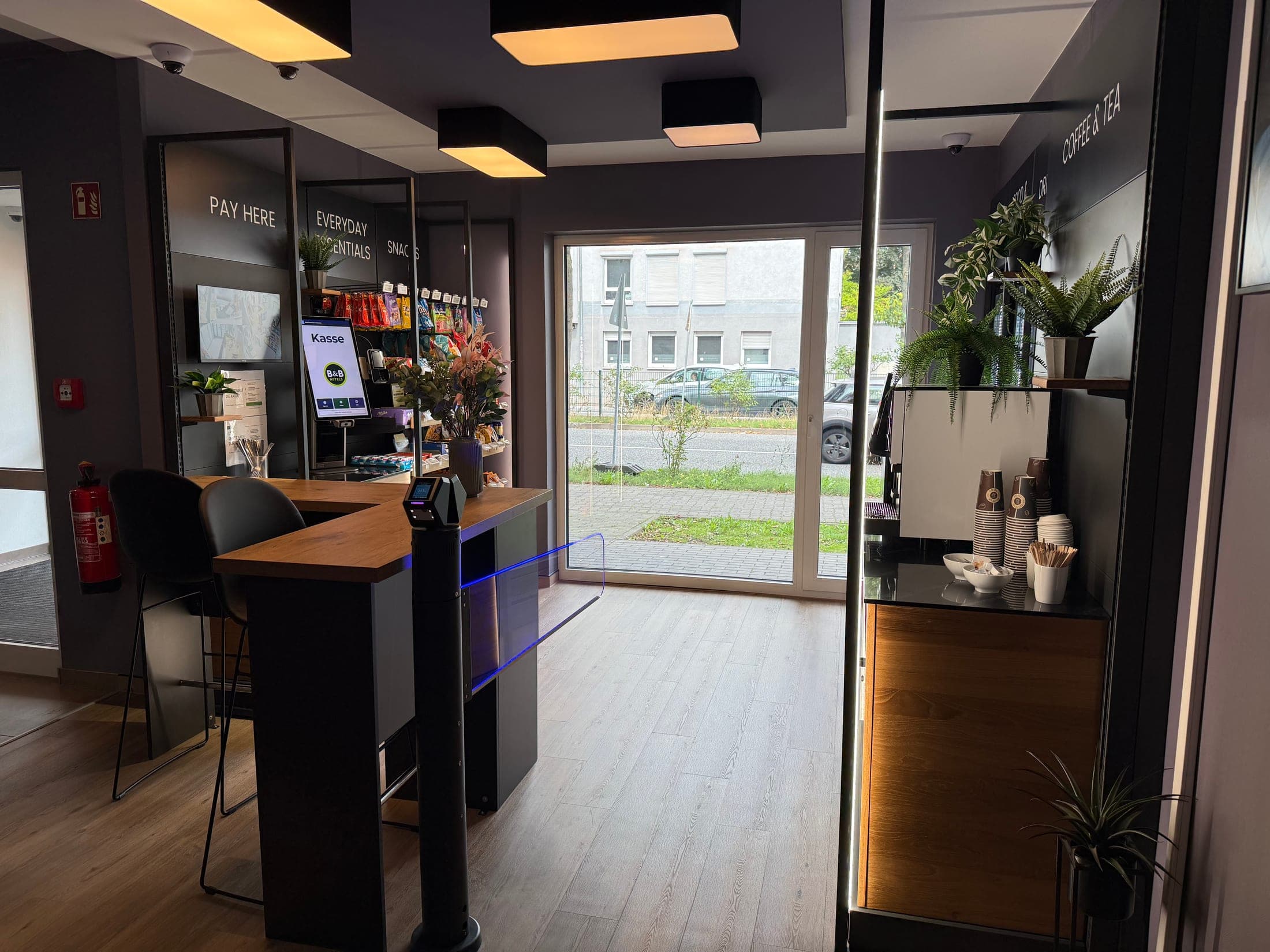 Blick durch den Verkaufsraum des Shops zur Glasfront — Self-Checkout-Kasse, Snackregale, Sitzplätze und Kaffeebereich