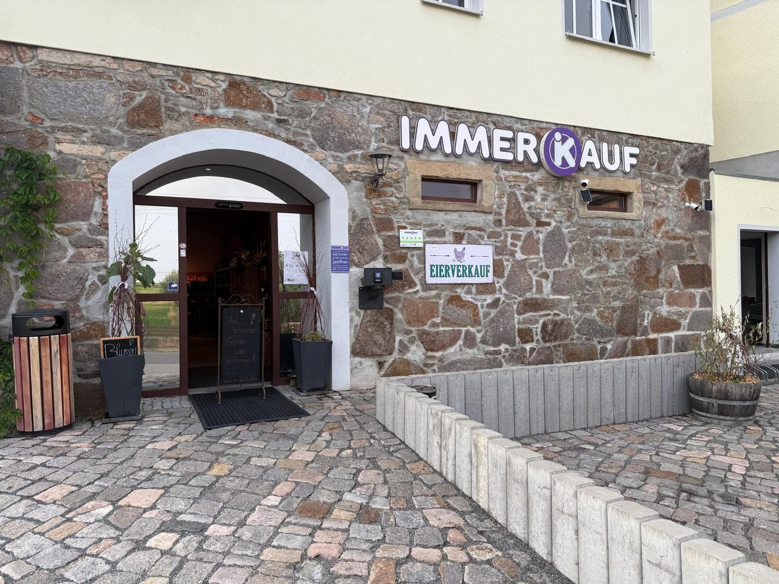 Eingang des Immer(K)auf Tag- und Nachtmarkts in Taupadel — 24 Stunden geöffnet, mit Überwachungskamera an der Natursteinfassade