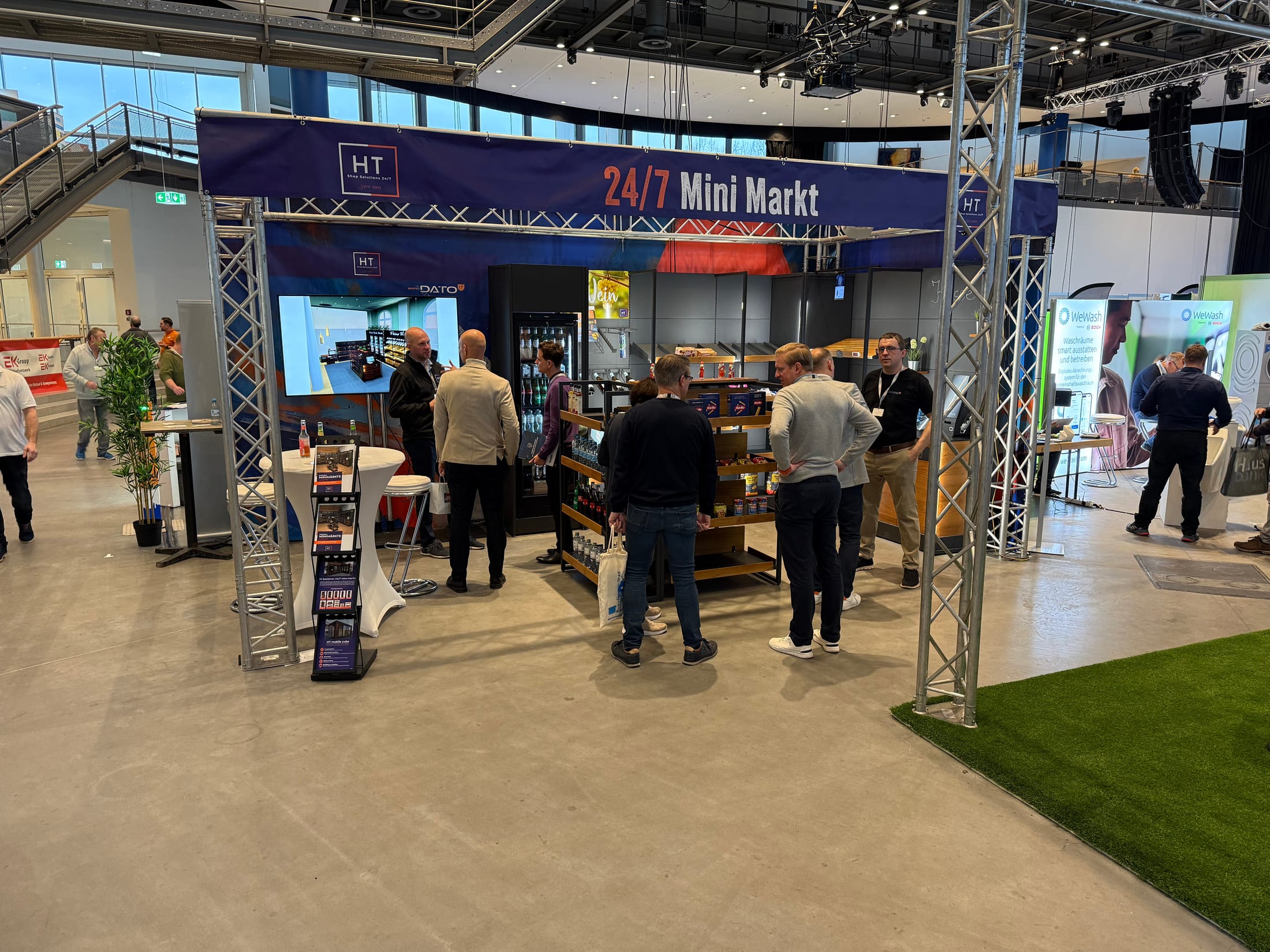 Besucher am 24/7-Mini-Markt-Stand auf der NCT 2025 in Lübeck — Regale mit Backwaren, Kühlschrank und Präsentationsbildschirm
